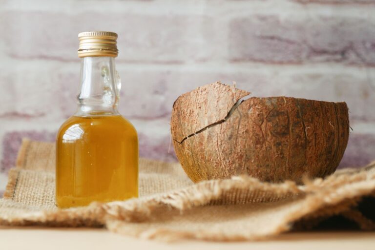 Bottle of coconut oil beside a cracked coconut shell on burlap fabric indoors.